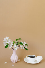 Beautiful branches of a blooming apple tree in a pink vase with a cup of tea and marshmallows on a beige background with a copy space