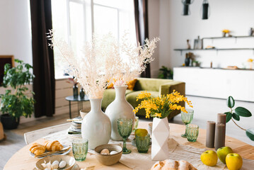 Festive Easter table with mimosa flowers in a vase and traditional food, eggs. The interior of a bright living room with a round set table