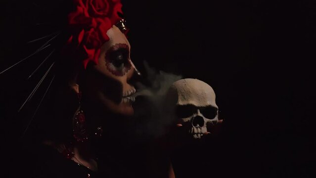 A woman with a pattern on her face in the style of the Goddess of Death with a sugar skull in front of her face on a black background blows white smoke on the scoop. Festival Day of the Dead