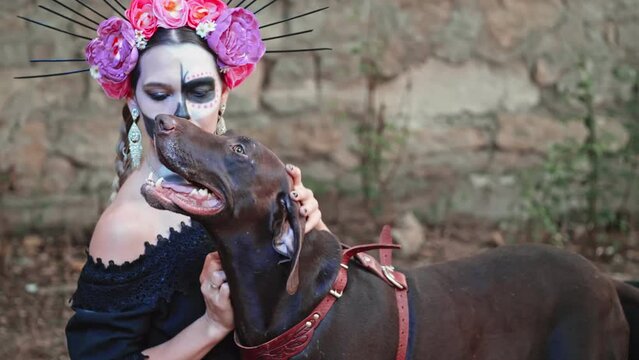 A young Woman with a design on the face and neck in the style of the Goddess of Death strokes a contented dog. Kindness and tenderness of the ancient holiday