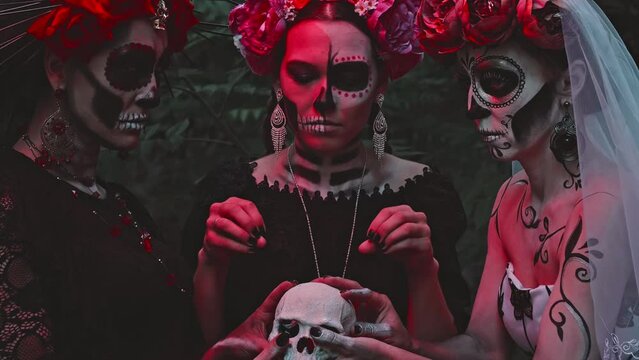 Three Young Women with a design on the face and neck in the style of the Goddess of Death or Katarina conjure over a sugar skull while holding it in their hands. 
