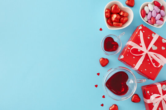 Valentines Day celebration concept. Top view photo of red gift boxes, heart shaped glass cup with drinking, confectionery chocolate candies and sprinkles on blue background with copy space.