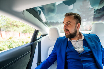 A caucasian man in a blue suit looks out the open window while sitting in the back seat of a car. Business class passenger. 