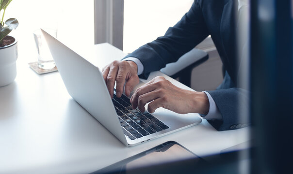Business man working on laptop computer at modern office. Businessman in black suit working and typing on laptop keyboard, surfing the internet with digital tablet on office table