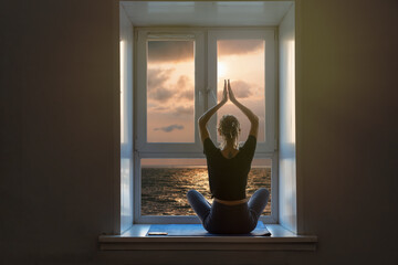Slim woman doing yoga meditation indoor at home, sea sunset view from  window