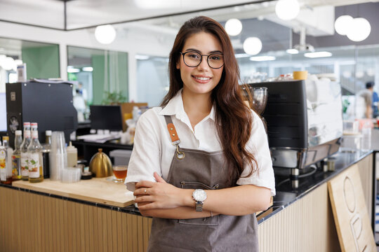 young beautiful barista woman is standing and cross arm with coffee shop background.