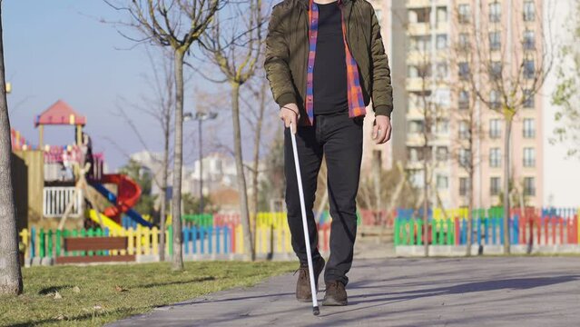 Blind man with white cane walking on the road.
Blind man walking on a path by the playground.
