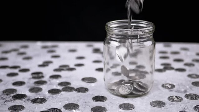 Silver Coins Fall into a Glass Jar against Background of Scattered Kopecks. Piggy bank. Stream of Ukrainian cents pours into a glass jar. Financial crisis, growth, budget savings, money accumulation.