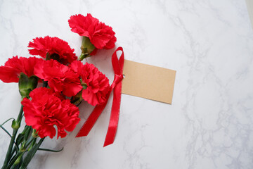 Red Carnation decoration with ribbon and blank message tag on white marble background. Carnation flower composition for Mother's day, Women's day and wedding celebration design. 