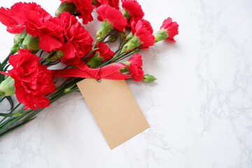 Red Carnation decoration with ribbon and blank message tag on white marble background. Carnation flower composition for Mother's day, Women's day and wedding celebration design. 