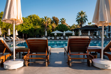 Empty deck chairs and white parasols arranged at poolside with lush trees and clear blue sky in the background at tourist resort
