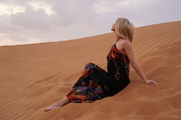 Beautiful blonde woman in a black dress is sitting on the hot sand in the desert