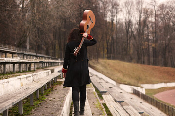 A woman in black walks with her back, with a guitar along a wooden bench at the old stadium on...