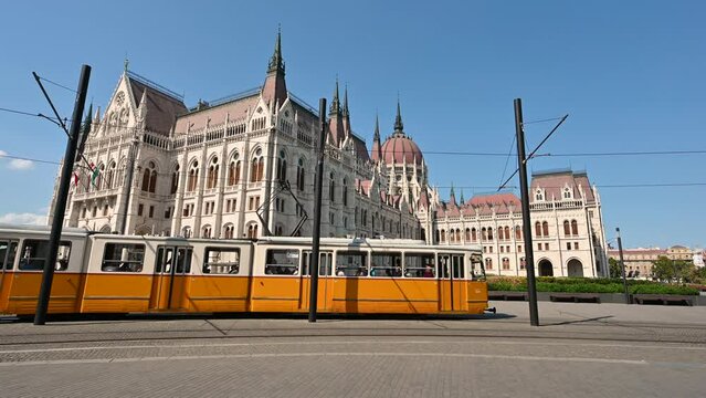 Budapest, Hungary, August 2022.Nice footage with iconic image of the city with wide angle lens. In the background of the parliament passes one of the orange vintage trams making its journey.