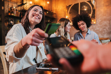 Happy young woman paying bill with a contactless credit card in a restaurant. Female smiling holding a creditcard and giving a payment transaction to the cashier. High quality photo - obrazy, fototapety, plakaty