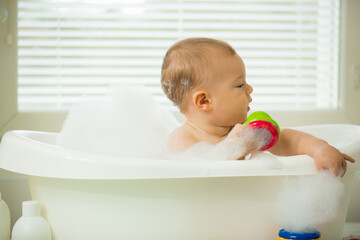 Cute little baby sitting in white bathtub with foam and soap bubbles. Taking bath and playing with toys. Baby hygiene. 