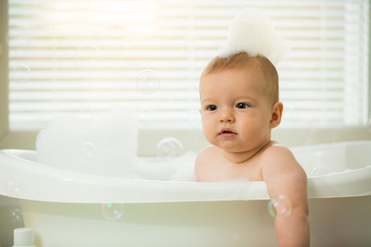Cute little baby sitting in white bathtub with foam and soap bubbles. Taking bath and playing with toys. Baby hygiene. 