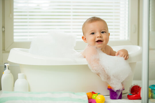 Cute little baby sitting in white bathtub with foam and soap bubbles. Taking bath and playing with toys. Baby hygiene. 