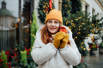 Woman holding a one Christmas gift outdoor in winter time. Snowfall. Fir-tree, garland backround. Concept of shopping, holidays and happiness Christmas.