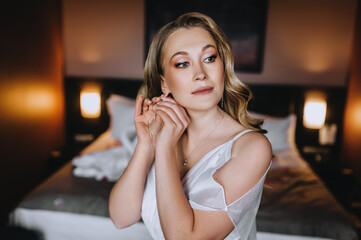 A beautiful curly blonde bride in a white dress straightens an earring on her ear while standing in the room. Wedding photography, close-up portrait.