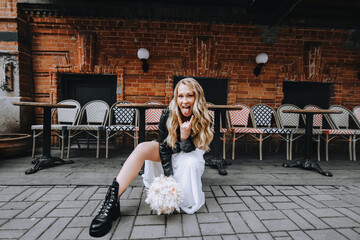 A beautiful curly, cool bride, biker, rock lover in a black leather jacket, white dress with a bouquet sits in the city against the backdrop of a brick wall. Wedding photography, portrait.