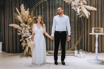 A stylish groom in a white shirt and a beautiful bride in a white dress stand holding hands in a...