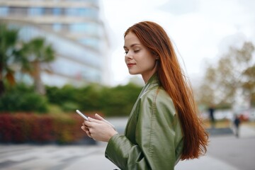 Fototapeta premium Woman with phone walking down the street in the city in fashionable clothes with red hair and a smile with teeth