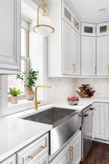 A kitchen sink detail shot with a gold pendant light, stainless steel apron sink, white cabinets,...