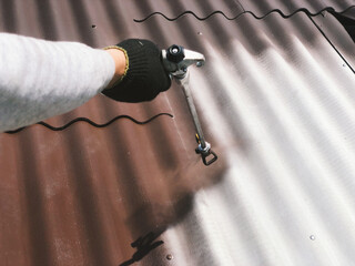 A worker holds a high-pressure spray gun in his hand and paints the roof of the house with brown paint