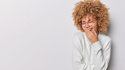 Fototapeta na wymiar Positive happy woman with curly hair covers mouth and hides emotions laughs silently wears transparent eyeglasses and casual white jumper poses against grey background blank space for your promotion