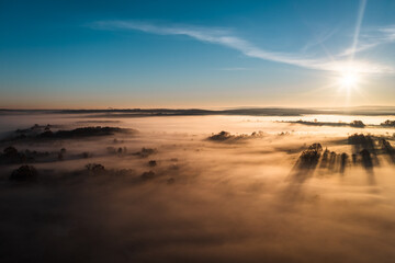 Flying over a village at dawn, bright sun on the horizon and fog over a small village.