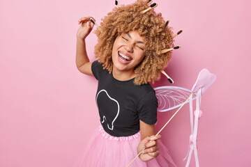Positive optimistic female tooth fairy dances carefree holds magic wand laughs happily wears black t shirt and pleated skirt tells how to care about your teeth isolated over pink background.