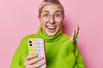 Joyful optimistic woman with short hair keeps hand raised up smiles broadly wears big transparent eyeglasses and green jumper holds mobile phone gets excellent news isolated over pink background - obrazy, fototapety, plakaty