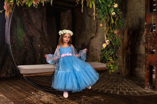 Little girl princess with blue eyes sits on fairy swing in dark mystery room, wearing art light blue dress with wreath of flowers on head, looking at camera. Fantasy art photo, fairy tale concept