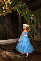 Obraz na płótnie Canvas Rear view little girl princess wearing art light blue dress with wreath of flowers on head posing in mystery room with fairy swing, looking at camera. Fantasy art photo, fairy tale concept. Copy space