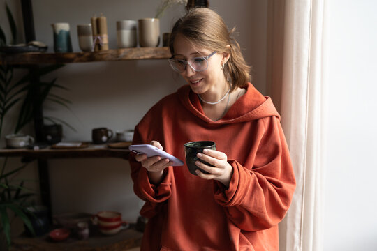 Young happy woman ceramic shop owner using phone and drinking tea, chatting with clients online. Female ceramist taking break while working in cozy pottery studio, potter resting at workplace