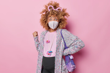 Shocked pregnant woman with curly hair clenches fist stares impressed at camera wears protective facial mask against coronavirus carries net bag isolated over pink wall. Pregnancy quarantine concept