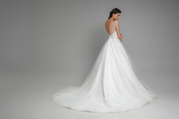 Fototapeta premium Portrait from the back of beautiful caucasian brunette bride in the beautiful white wedding dress standing in the grey studio. Gorgeous beautiful female on a isolated background.