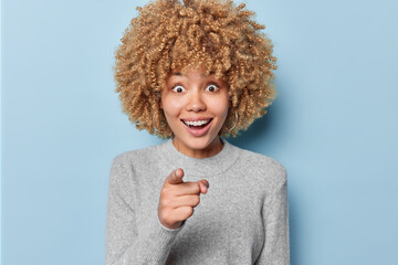 Happy surprised European woman with curly blonde hair points index finger directly at camera selects someone looks with great wonder at camera isolated over blue background. Wow thats awesome