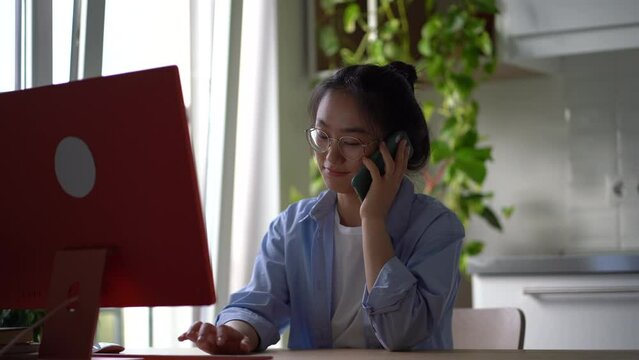 Korean girl remote employee having phone conversation while working on computer from home, talking with friend and typing on keyboard, sitting at desk in kitchen. Remote work distractions