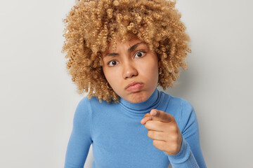 Portrait of serious curly haired woman points index finger at camera blames you and feels angry dressed in blue turtleneck isolated over grey background. Displeased female model indicates at camera