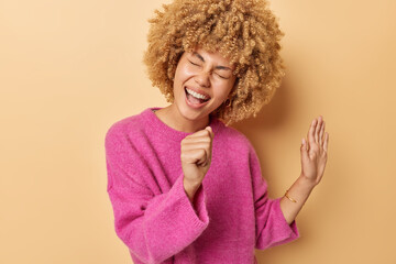 Positive carefree woman with curly hair keeps hand near mouth as if microphone dressed in casual...