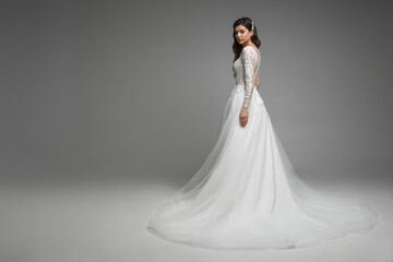 Full length of the beautiful caucasian brunette bride in the beautiful white wedding dress with pearls and embroidery standing, posing with hands in the grey studio.