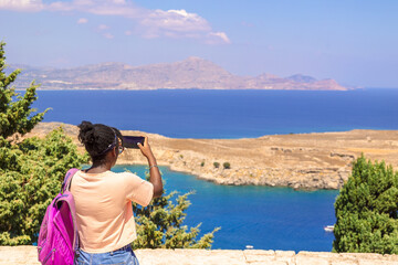Fototapeta na wymiar Happy girl enjoys view of coast of ancient old town of city Lindos, Greece. Woman in taking photo on phone of colorful panoramic views of coastline Rhodes in Aegean Sea. Beautiful landscape