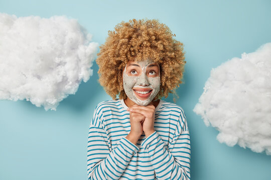 Portrait of dreamy curly haired woman keeps hands under chin focused above applies facial clay mask for skin care dressed in long sleeved striped jumper isolated on blue background white clouds above
