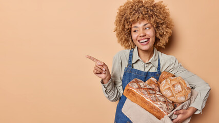 Positive curly housewife bakes fresh bread at home indicates index finger aside and shows place for your promotional content smiles broadly wears shirt and apron isolated over beige background