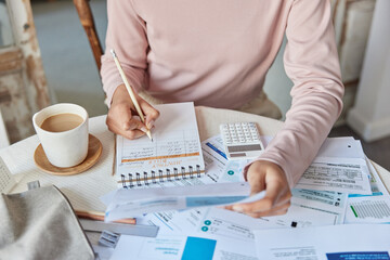 Cropped shot of unknown woman poses at home office makes calculations of utility payments writes down notes in spiral notebook surrounded by paper bills and cup of coffee calculates domestic expenses - obrazy, fototapety, plakaty