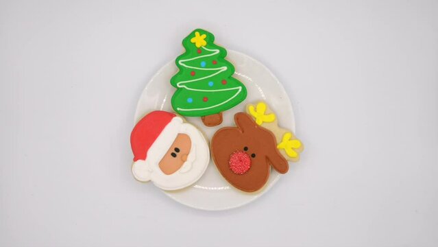 HD video of three fancy decorated Christmas cookies on a round porcelain plate from above, caucasian hand reaches in and adds three presents then removes one cookie.
