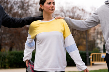 Serious young female athlete in colorful sweatshirt standing between sportsmen while stretching leg in park