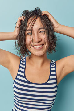 Happy Asian woman has wet hair after douching looks gladfully aside keeps hands on head wears striped t shirt takes care of herself isolated over blue background. Beauty and hygiene concept.